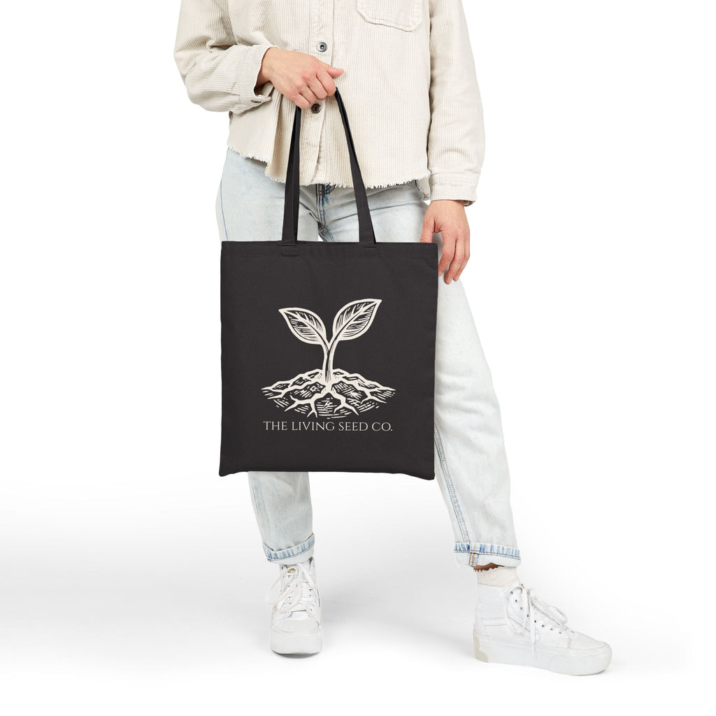 Person carrying The Original Canvas Tote in Black, showing full size and 20-inch handles with etching-style sprouting seed graphic and The Living Seed Co. wordmark in parchment ink