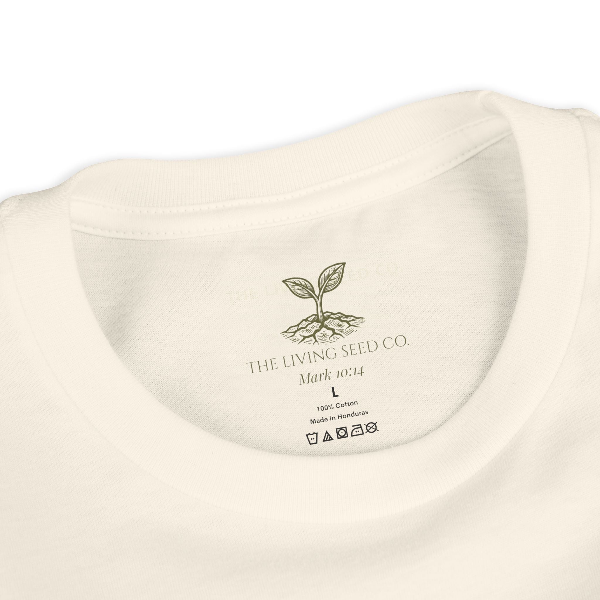 The Original Youth Tee in Natural — neck label closeup — custom printed label featuring The Living Seed Co. logo, Mark 10:14, size, fabric content, and care icons. 100% cotton, made in Honduras.
