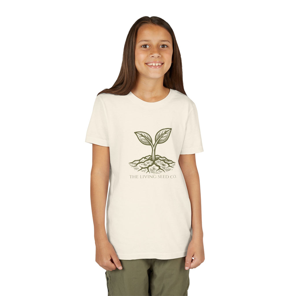 The Original Youth Tee in Natural — lifestyle shot — girl wearing cream youth t-shirt featuring The Living Seed Co. etching-style sprouting seed graphic in olive ink with wordmark below. Bella+Canvas 3001Y.