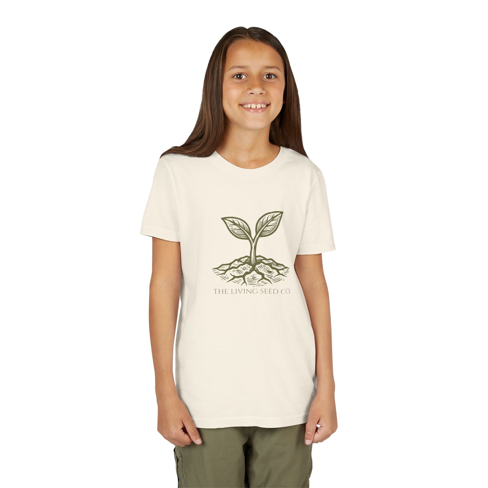 The Original Youth Tee in Natural — lifestyle shot — girl wearing cream youth t-shirt featuring The Living Seed Co. etching-style sprouting seed graphic in olive ink with wordmark below. Bella+Canvas 3001Y.