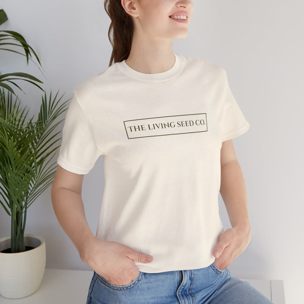 The Original Tee in Natural — front view — woman wearing cream unisex t-shirt featuring The Living Seed Co. boxed wordmark on chest in olive ink. Bella+Canvas 3001.