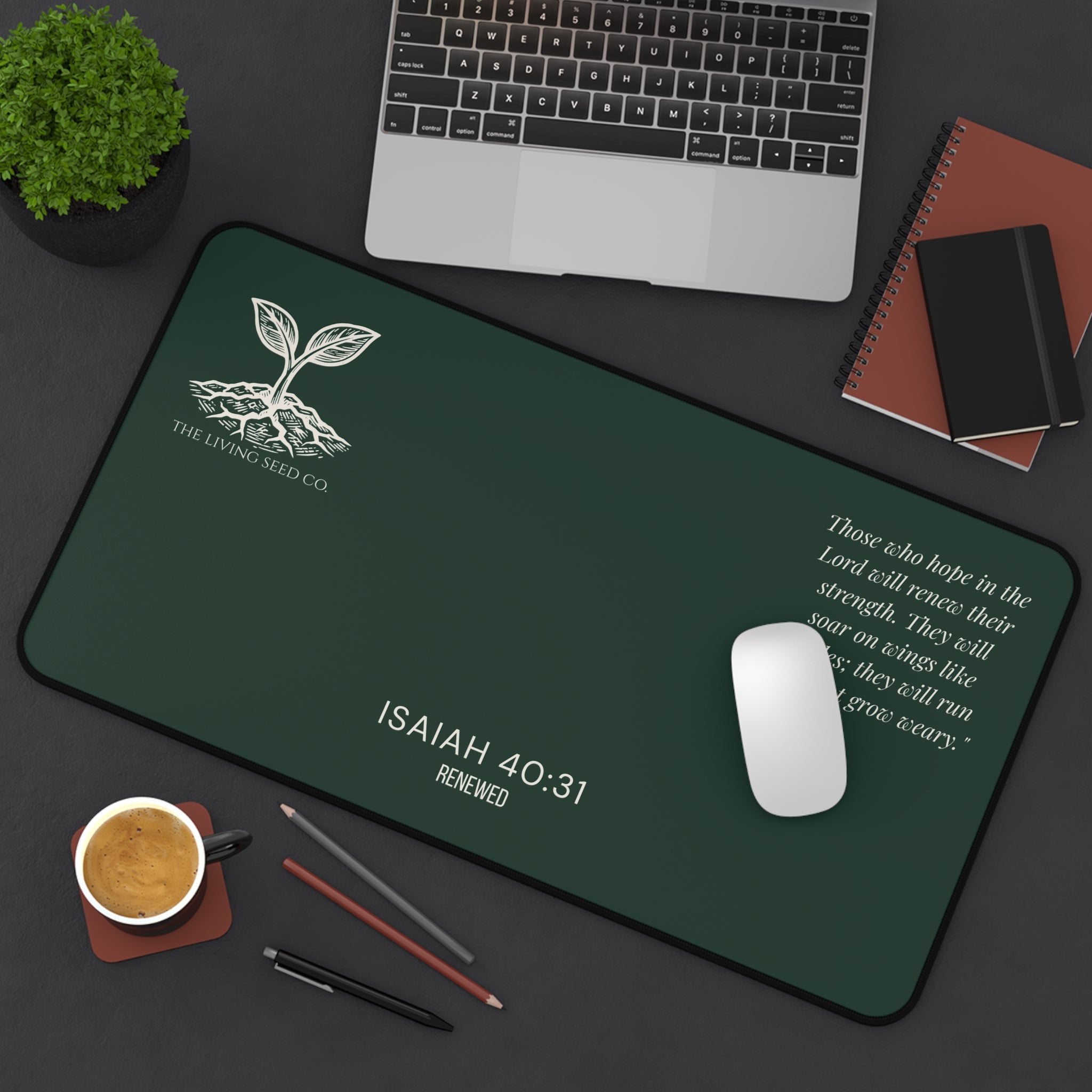 Scripture Series RENEWED Desk Mat in Forest Deep 12x22 — lifestyle shot — forest green desk mat on a dark workspace with laptop, coffee, and stationery featuring The Living Seed Co. seedling logo, Isaiah 40:31, RENEWED wordmark, and full Isaiah 40:31 verse in parchment ink.