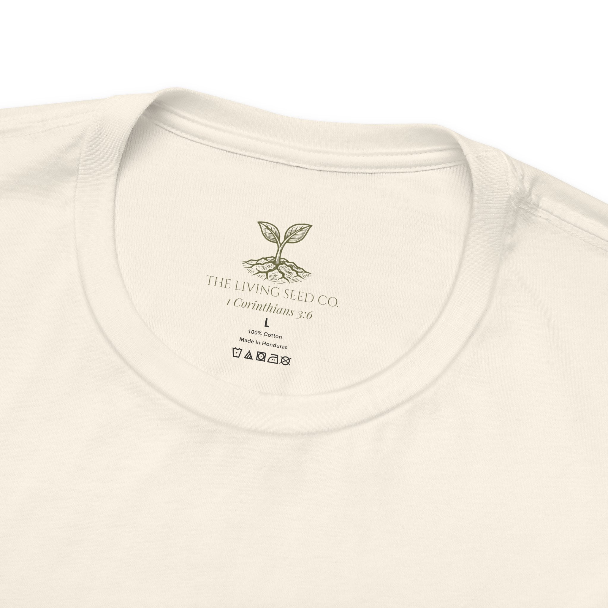 The Original Tee — neck label closeup — custom printed label featuring The Living Seed Co. logo, 1 Corinthians 3:6, size, fabric content, and care icons. 100% cotton, made in Honduras.