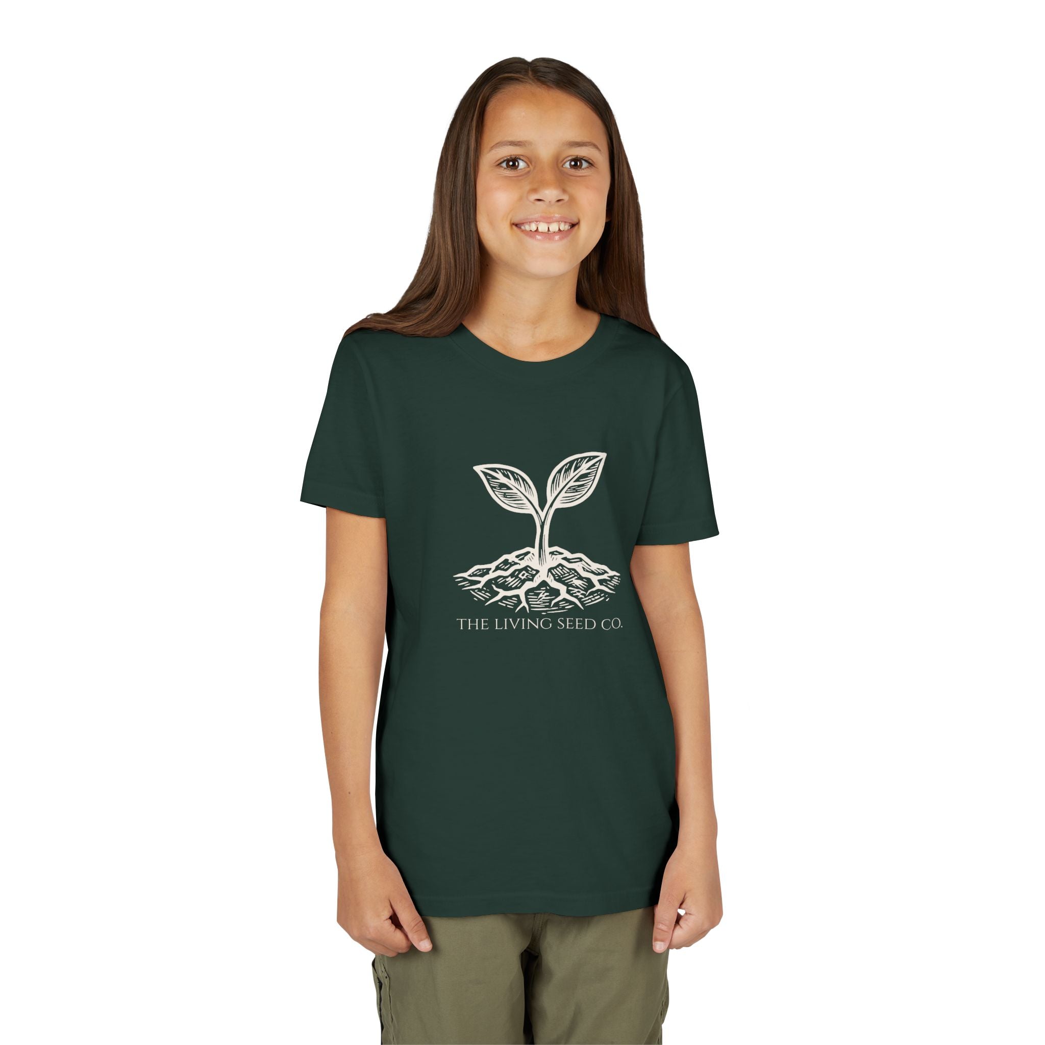 The Original Youth Tee in Forest — lifestyle shot — girl wearing dark green youth t-shirt featuring The Living Seed Co. etching-style sprouting seed graphic in parchment ink with wordmark below. Bella+Canvas 3001Y.