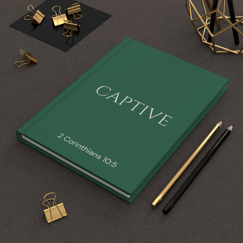 Scripture Series CAPTIVE Journal in Garden Green — lifestyle shot — garden green hardcover journal featuring CAPTIVE in parchment lettering with 2 Corinthians 10:5 reference, styled on a desk with gold stationery accessories.