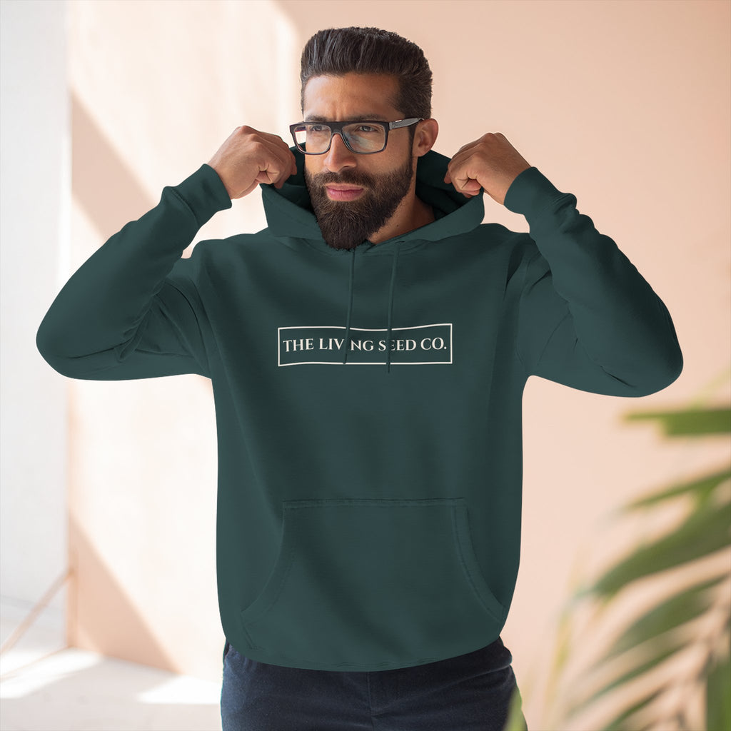 The Original Hoodie (Lane Seven) in Forest — lifestyle shot — man wearing forest green pullover hoodie featuring The Living Seed Co. boxed wordmark on chest in parchment ink. Lane Seven LS14001.