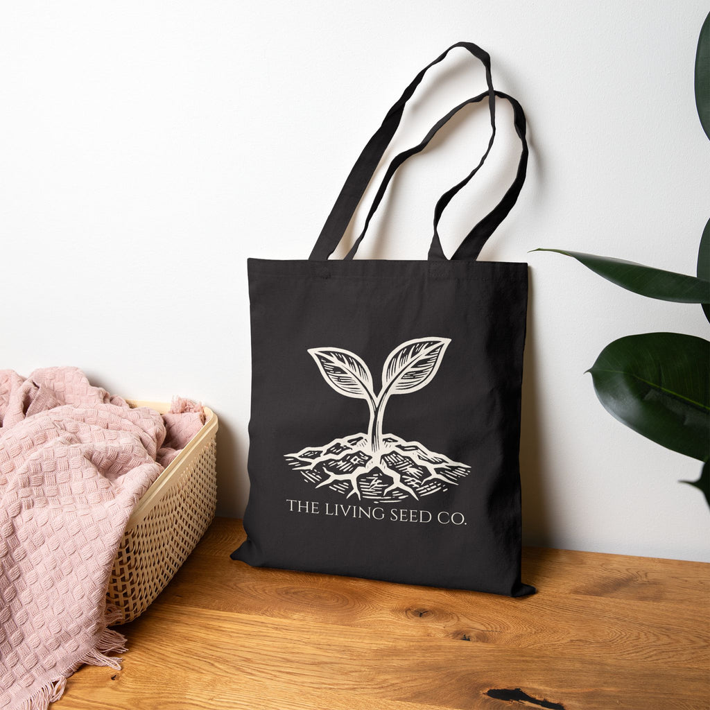 The Original Canvas Tote in Black standing on a wooden surface featuring an etching-style sprouting seed graphic and The Living Seed Co. wordmark in parchment ink