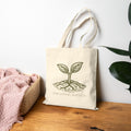 The Original Canvas Tote in Natural standing on a wooden surface featuring an etching-style sprouting seed graphic and The Living Seed Co. wordmark in olive ink