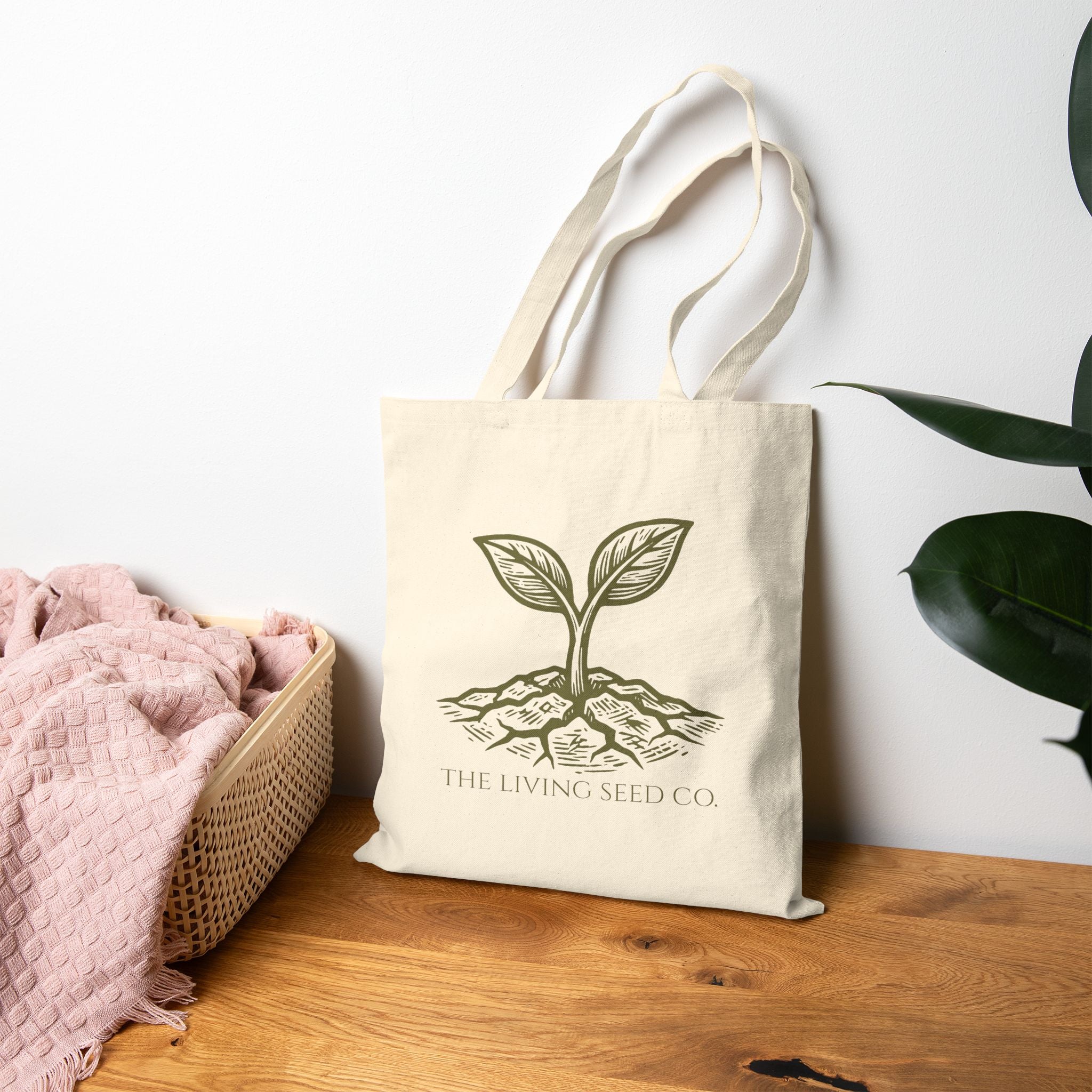 The Original Canvas Tote in Natural standing on a wooden surface featuring an etching-style sprouting seed graphic and The Living Seed Co. wordmark in olive ink