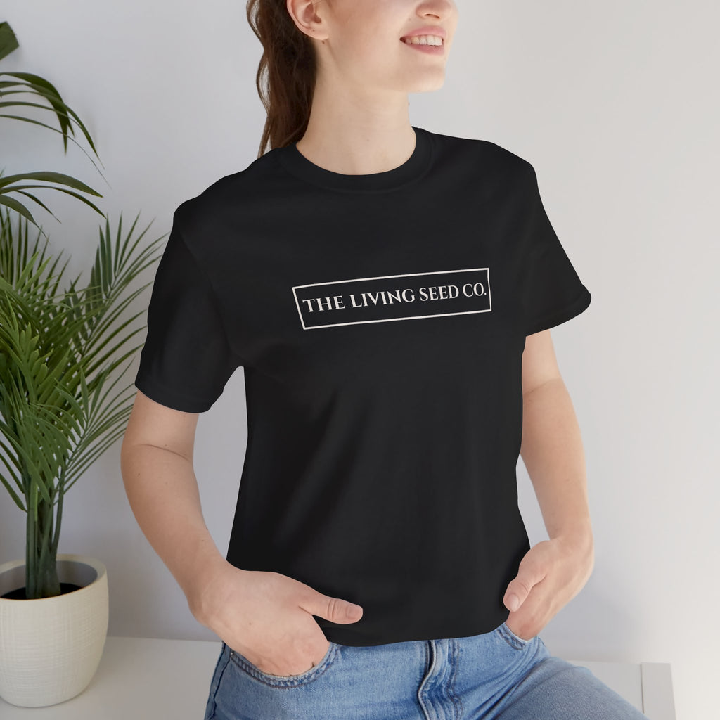 The Original Tee in Black — front view — woman wearing black unisex t-shirt featuring The Living Seed Co. boxed wordmark on chest in parchment ink. Bella+Canvas 3001.