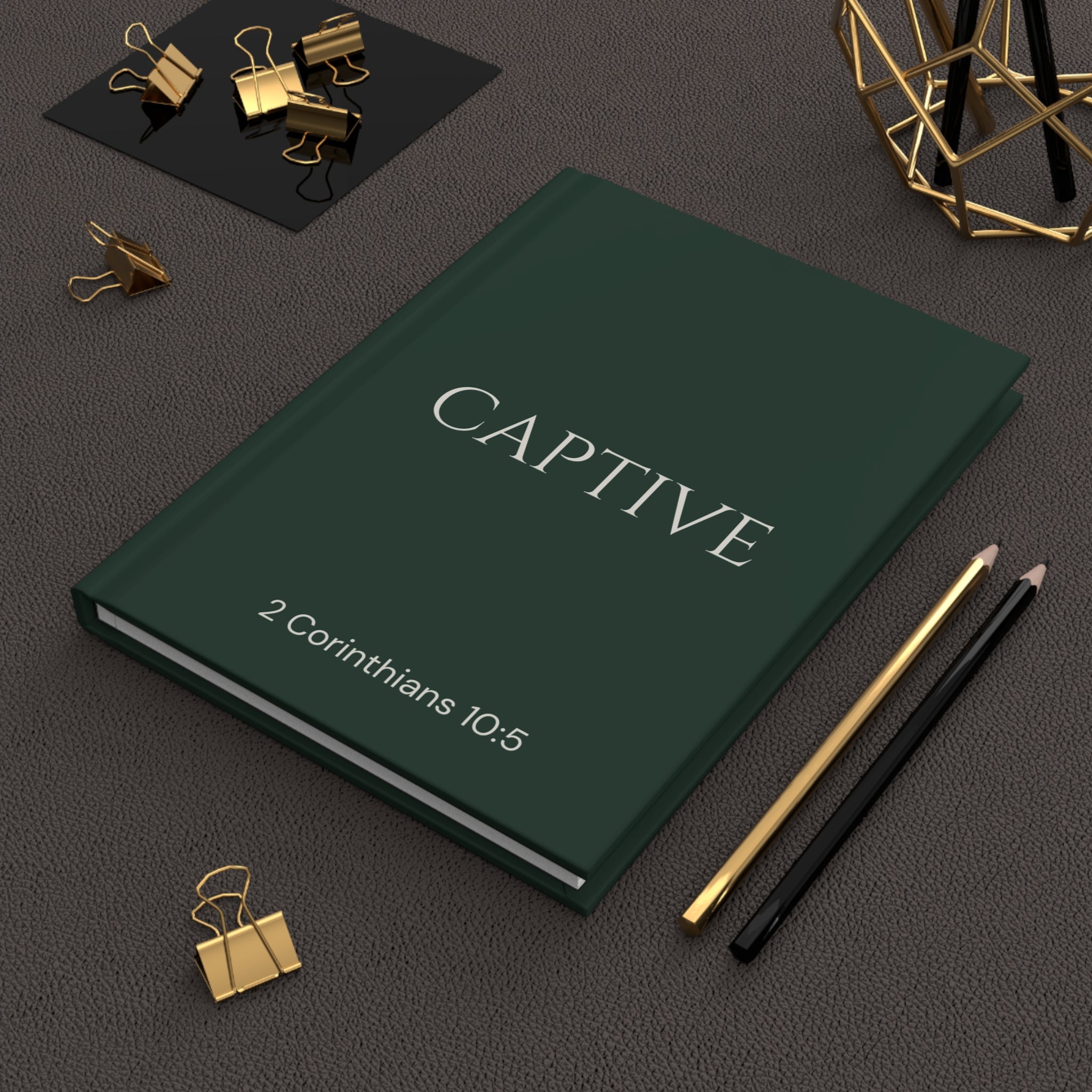 Scripture Series CAPTIVE Journal in Forest Deep — lifestyle shot — deep green hardcover journal featuring CAPTIVE in parchment lettering with 2 Corinthians 10:5 reference, styled on a desk with gold stationery accessories.