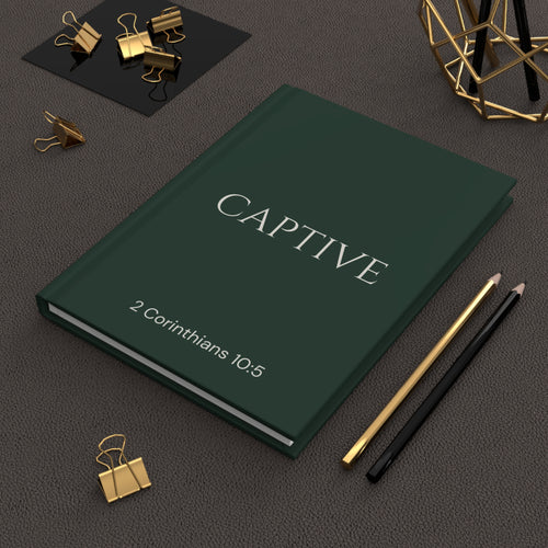 Scripture Series CAPTIVE Journal in Forest Deep — lifestyle shot — deep green hardcover journal featuring CAPTIVE in parchment lettering with 2 Corinthians 10:5 reference, styled on a desk with gold stationery accessories.