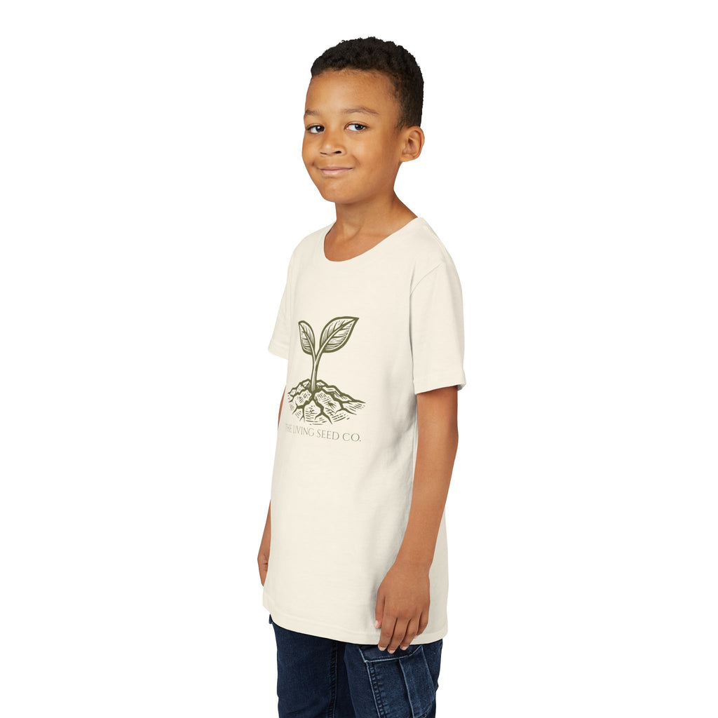 The Original Youth Tee in Natural — lifestyle shot — boy wearing cream youth t-shirt featuring The Living Seed Co. etching-style sprouting seed graphic in olive ink with wordmark below. Bella+Canvas 3001Y.