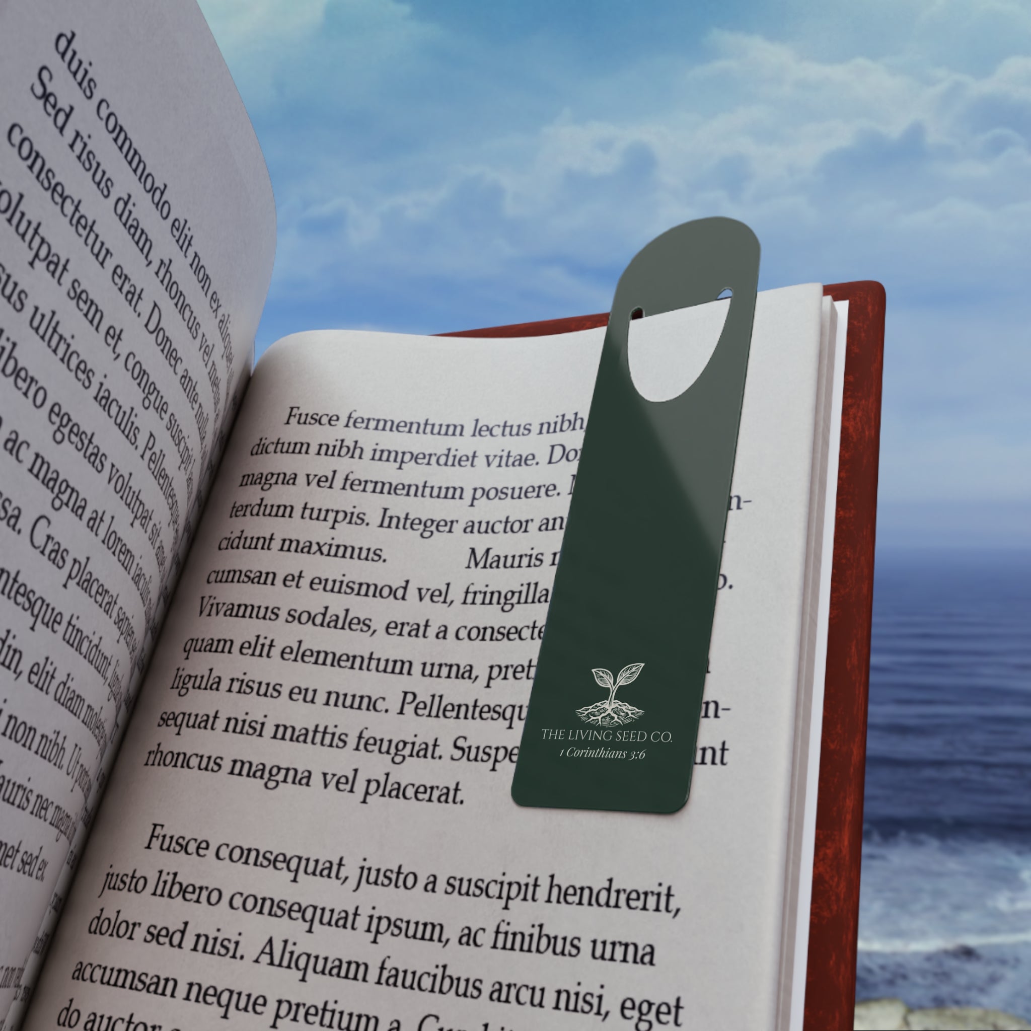 Forest Deep aluminum bookmark resting in an open book with an ocean view in the background, featuring The Living Seed Co. logo and 1 Corinthians 3:6