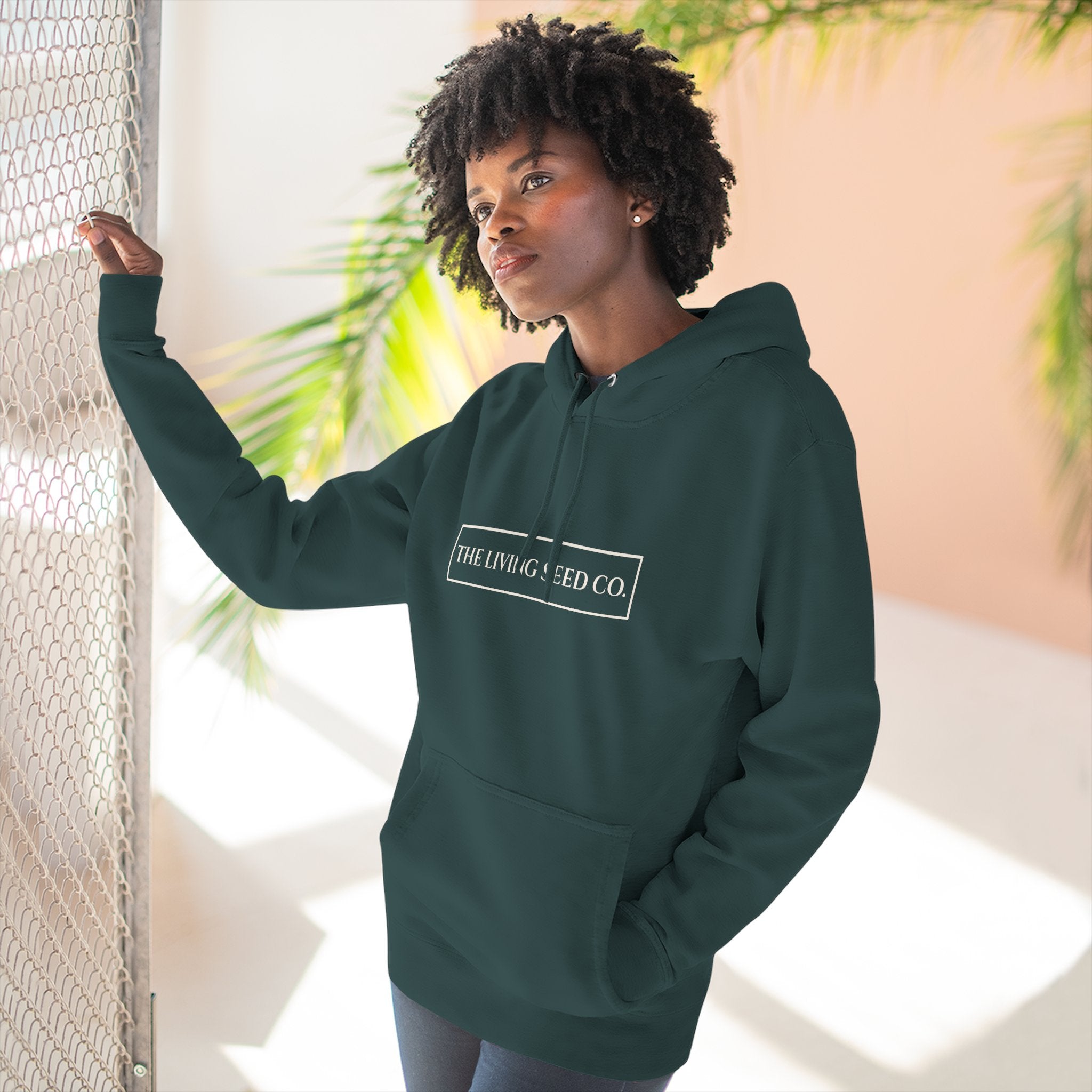 The Original Hoodie (Lane Seven) in Forest — lifestyle shot — woman wearing forest green pullover hoodie featuring The Living Seed Co. boxed wordmark on chest in parchment ink. Lane Seven LS14001.