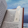 Parchment aluminum bookmark resting in an open book with an ocean view in the background, featuring The Living Seed Co. logo and 1 Corinthians 3:6