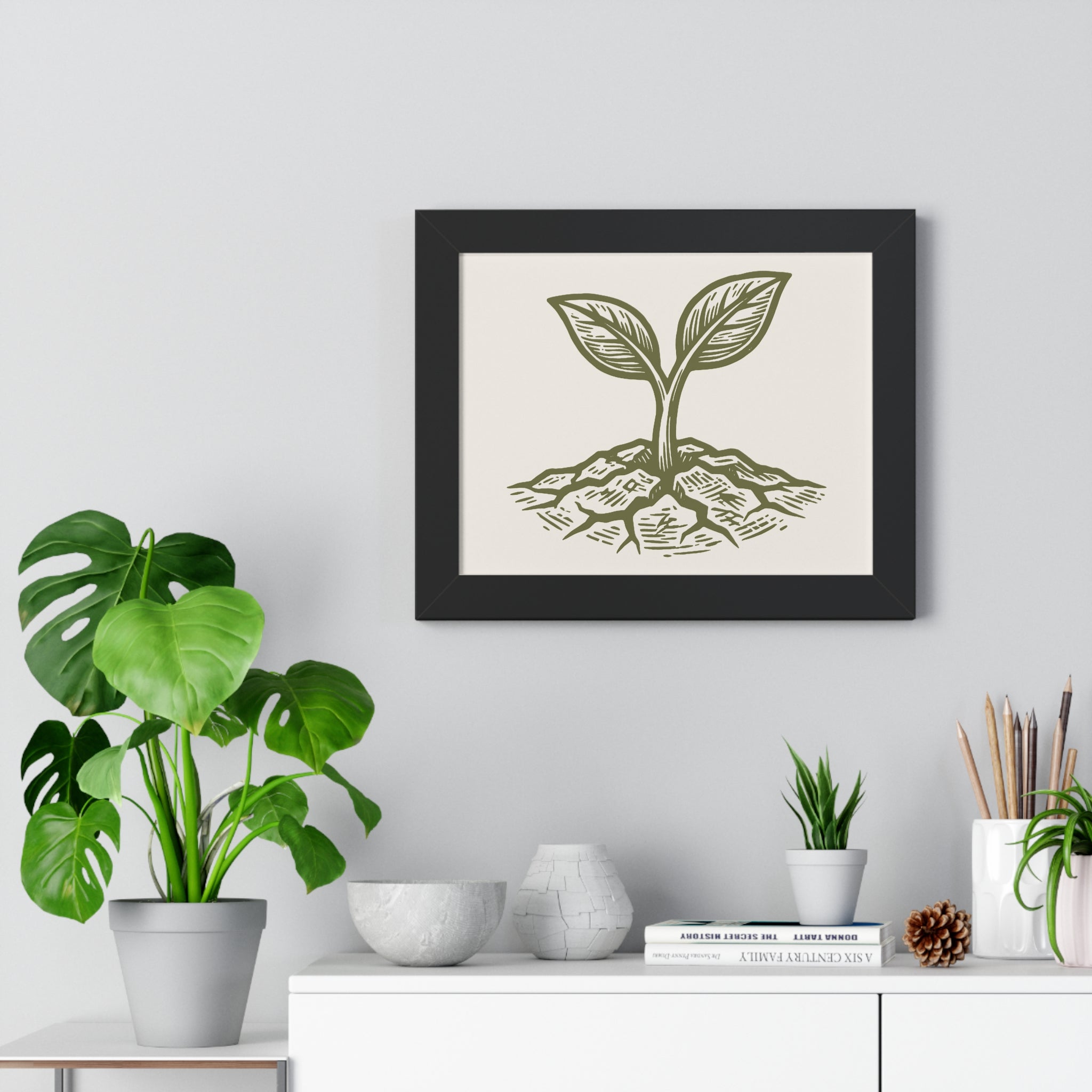 The Original Framed Poster in Black hanging on a gray wall above a shelf with houseplants, featuring an etching-style sprouting seed illustration on a Parchment background