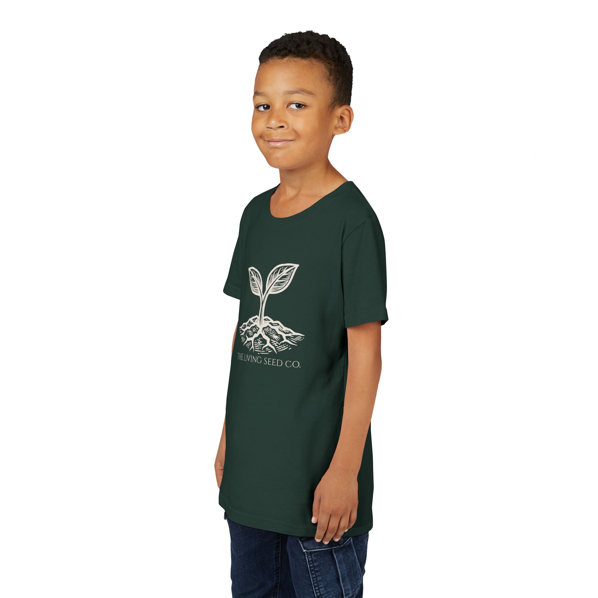 The Original Youth Tee in Forest — lifestyle shot — boy wearing dark green youth t-shirt featuring The Living Seed Co. etching-style sprouting seed graphic in parchment ink with wordmark below. Bella+Canvas 3001Y.