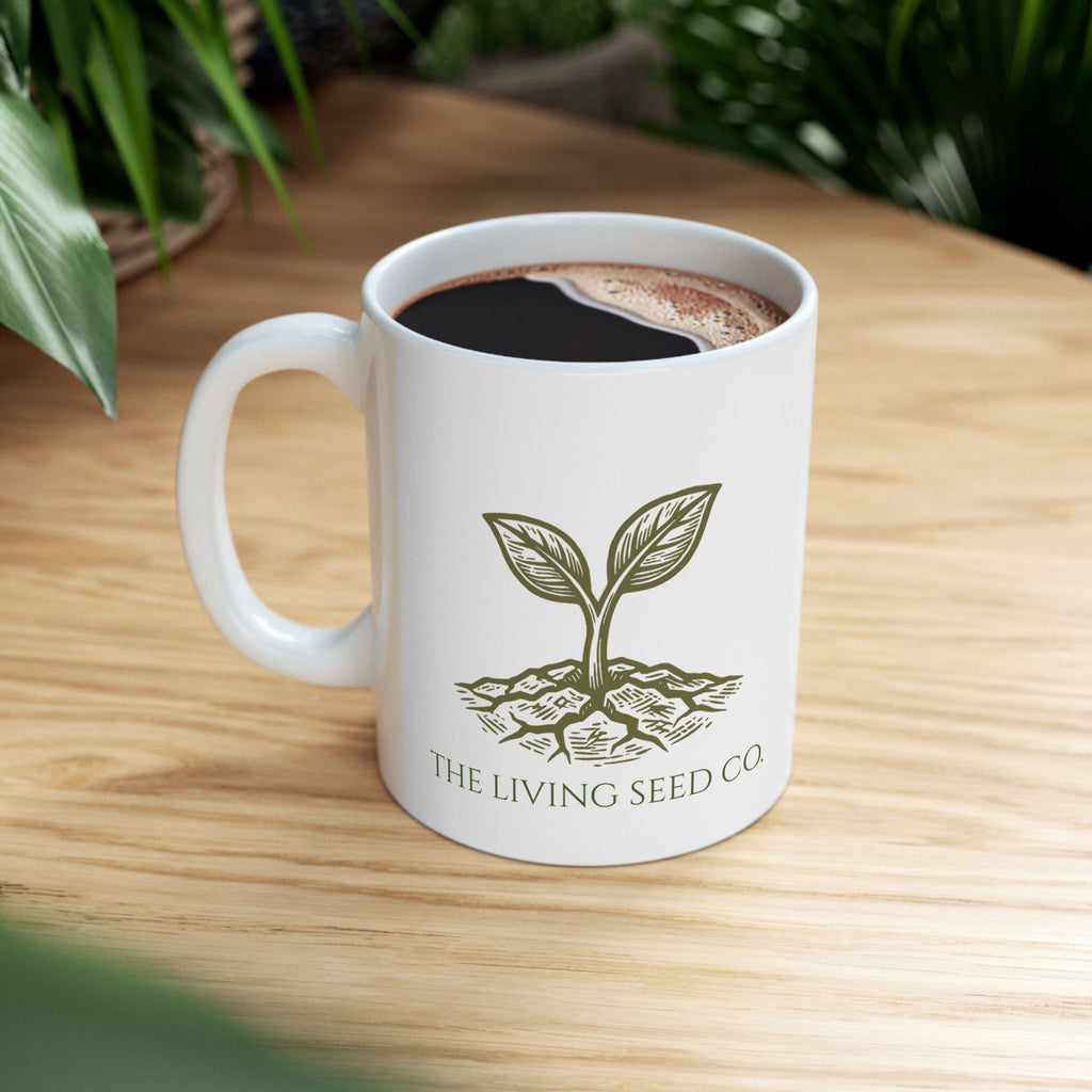 The Original Mug 11oz — lifestyle shot — white ceramic mug filled with coffee resting on a wood surface featuring The Living Seed Co. etching-style sprouting seed graphic in olive ink with wordmark below.