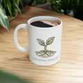 The Original Mug 11oz — lifestyle shot — white ceramic mug filled with coffee resting on a wood surface featuring The Living Seed Co. etching-style sprouting seed graphic in olive ink with wordmark below.