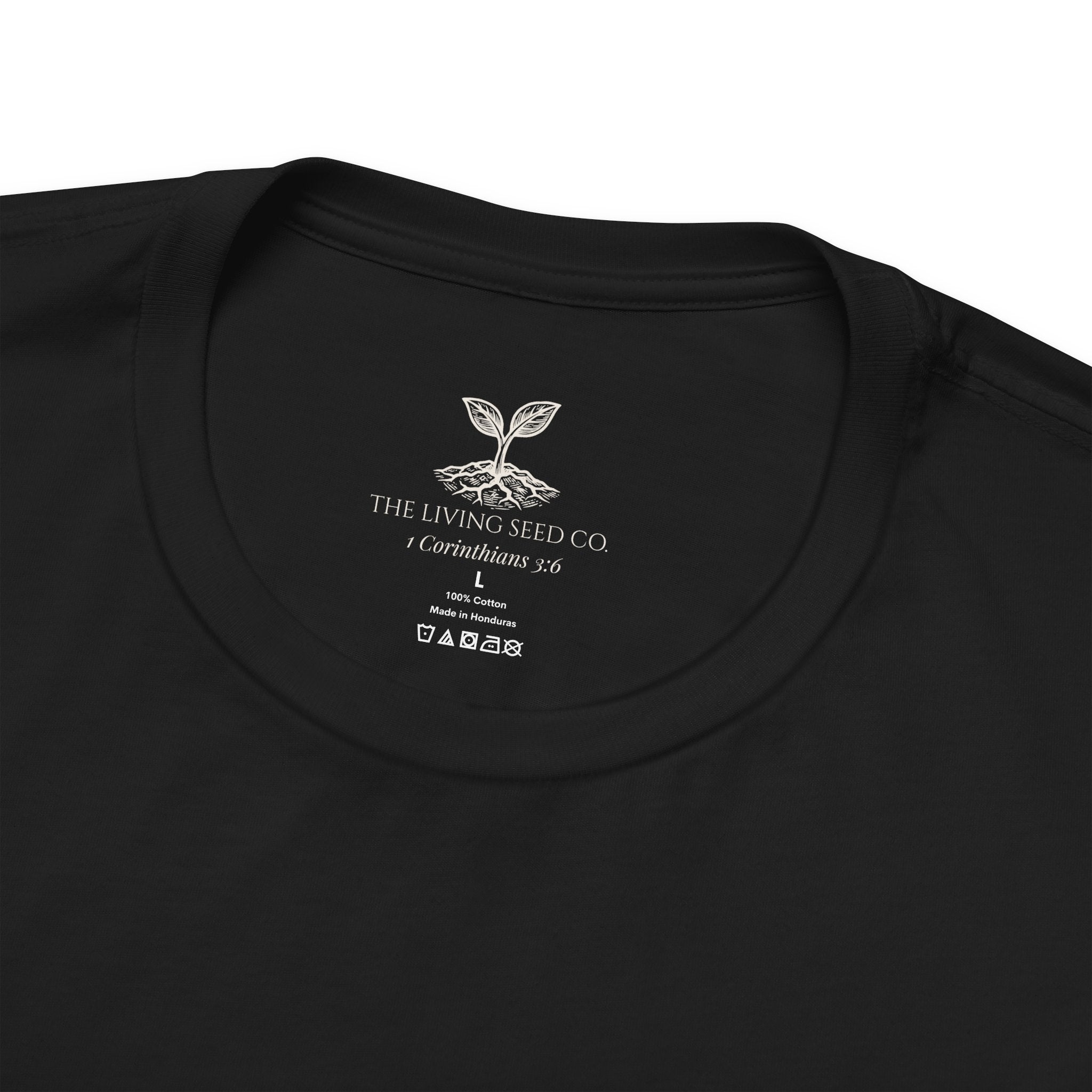 The Original Tee in Black — neck label closeup — custom printed label featuring The Living Seed Co. logo, 1 Corinthians 3:6, size, fabric content, and care icons. 100% cotton, made in Honduras.