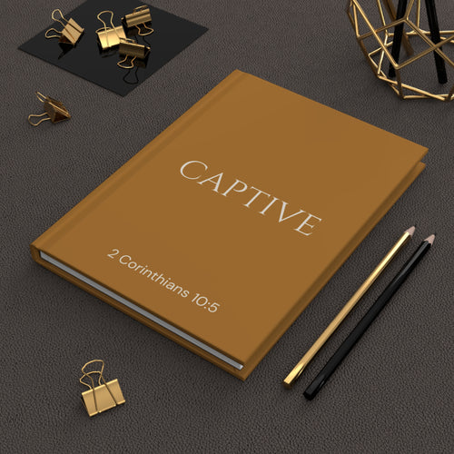 Scripture Series CAPTIVE Journal in Harvest Gold — hardcover journal featuring CAPTIVE in parchment lettering with 2 Corinthians 10:5 reference, styled on a desk with gold stationery accessories.