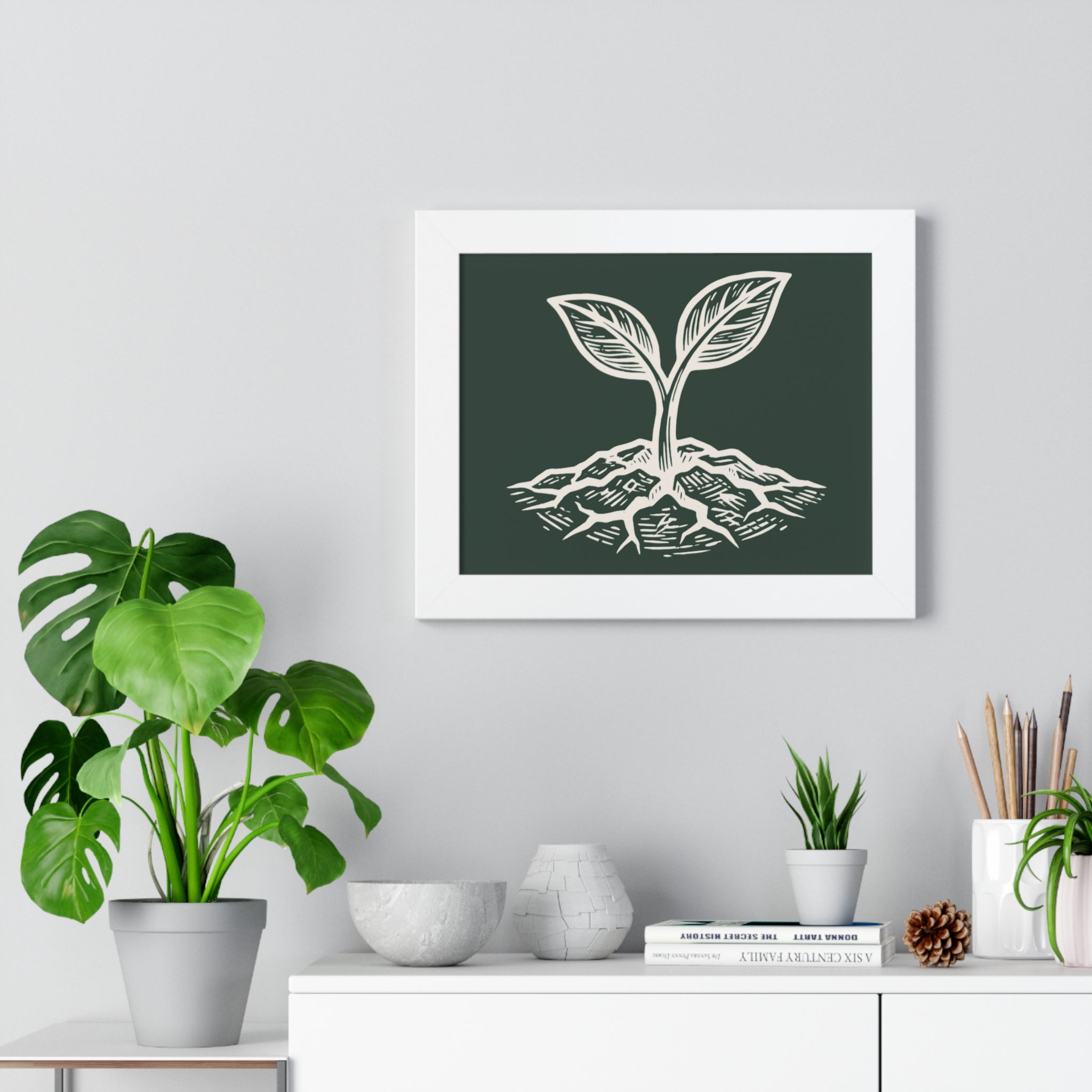 The Original Framed Poster in White hanging on a gray wall above a shelf with houseplants, featuring an etching-style sprouting seed illustration on a Forest Deep background