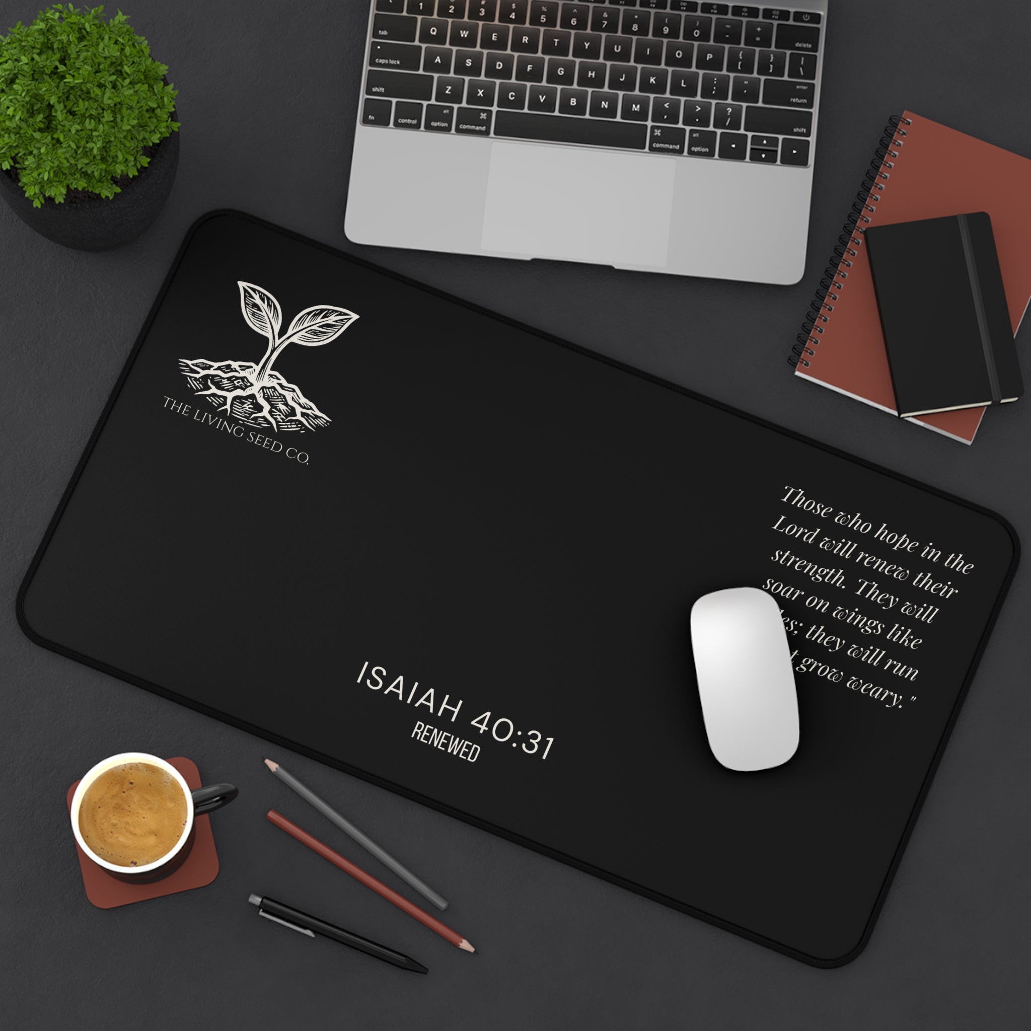 Scripture Series RENEWED Desk Mat in Black 12x22 — lifestyle shot — forest green desk mat on a dark workspace with laptop, coffee, and stationery featuring The Living Seed Co. seedling logo, Isaiah 40:31, RENEWED wordmark, and full Isaiah 40:31 verse in parchment ink.