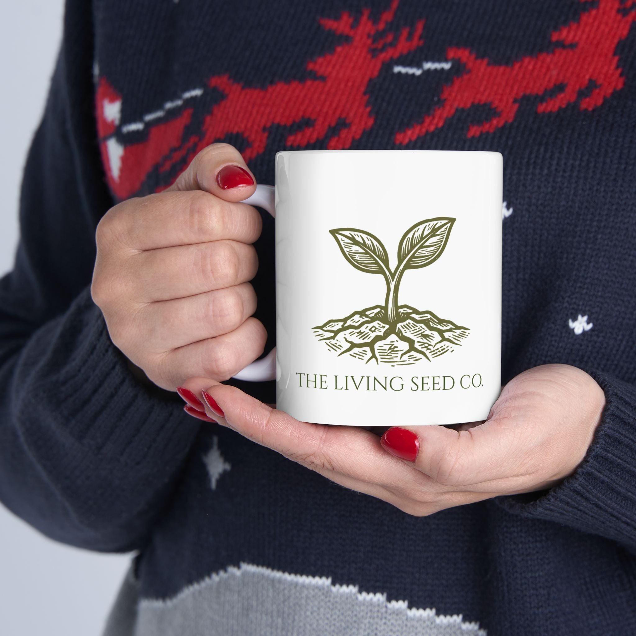 The Original Mug 11oz — lifestyle shot — woman holding white ceramic mug featuring The Living Seed Co. etching-style sprouting seed graphic in olive ink with wordmark below.