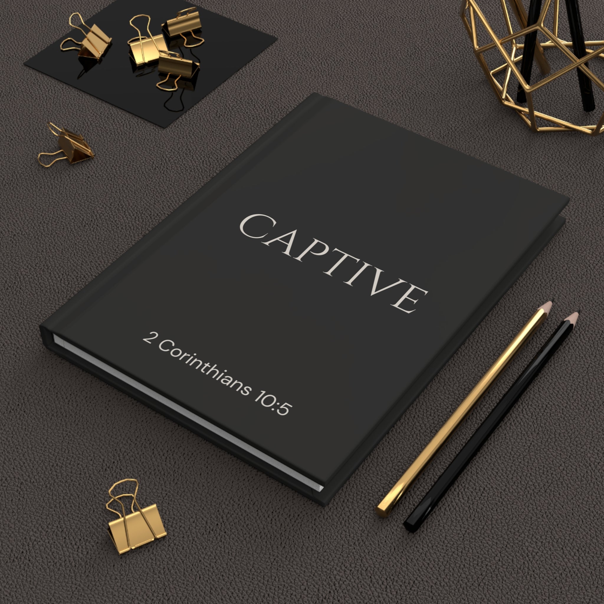 Scripture Series CAPTIVE Journal in Midnight Black — lifestyle shot — black hardcover journal featuring CAPTIVE in parchment lettering with 2 Corinthians 10:5 reference, styled on a desk with gold stationery accessories.
