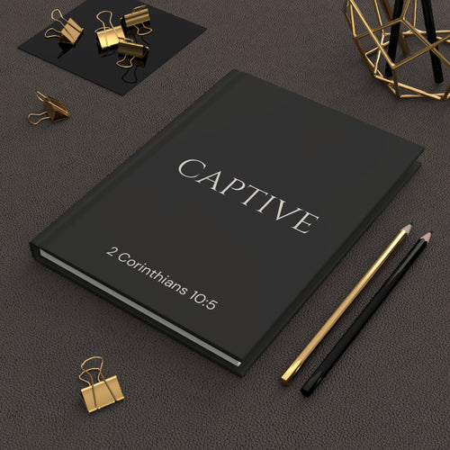 Scripture Series CAPTIVE Journal in Midnight Black — lifestyle shot — black hardcover journal featuring CAPTIVE in parchment lettering with 2 Corinthians 10:5 reference, styled on a desk with gold stationery accessories.