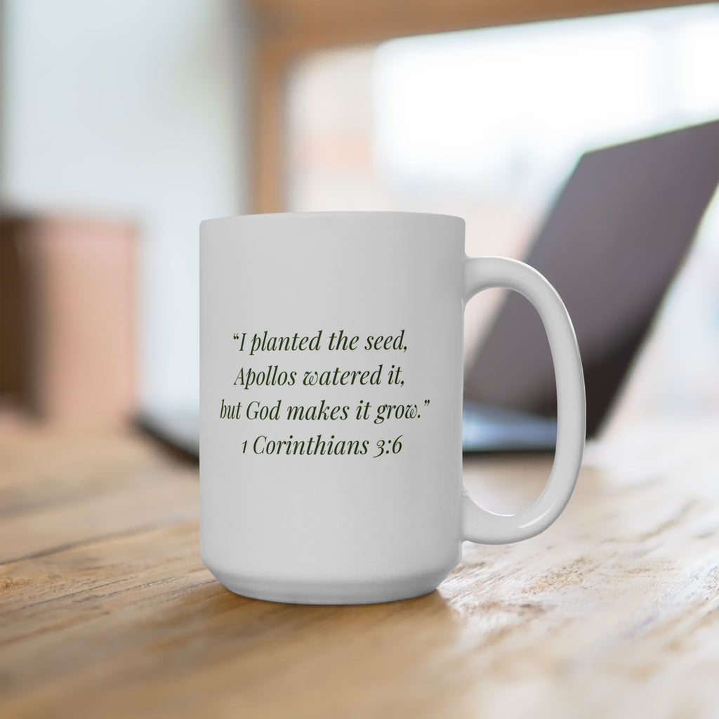 The Original Mug 15oz — scripture side lifestyle shot — white ceramic mug on a wood desk featuring 1 Corinthians 3:6 in olive ink: I planted the seed, Apollos watered it, but God makes it grow.