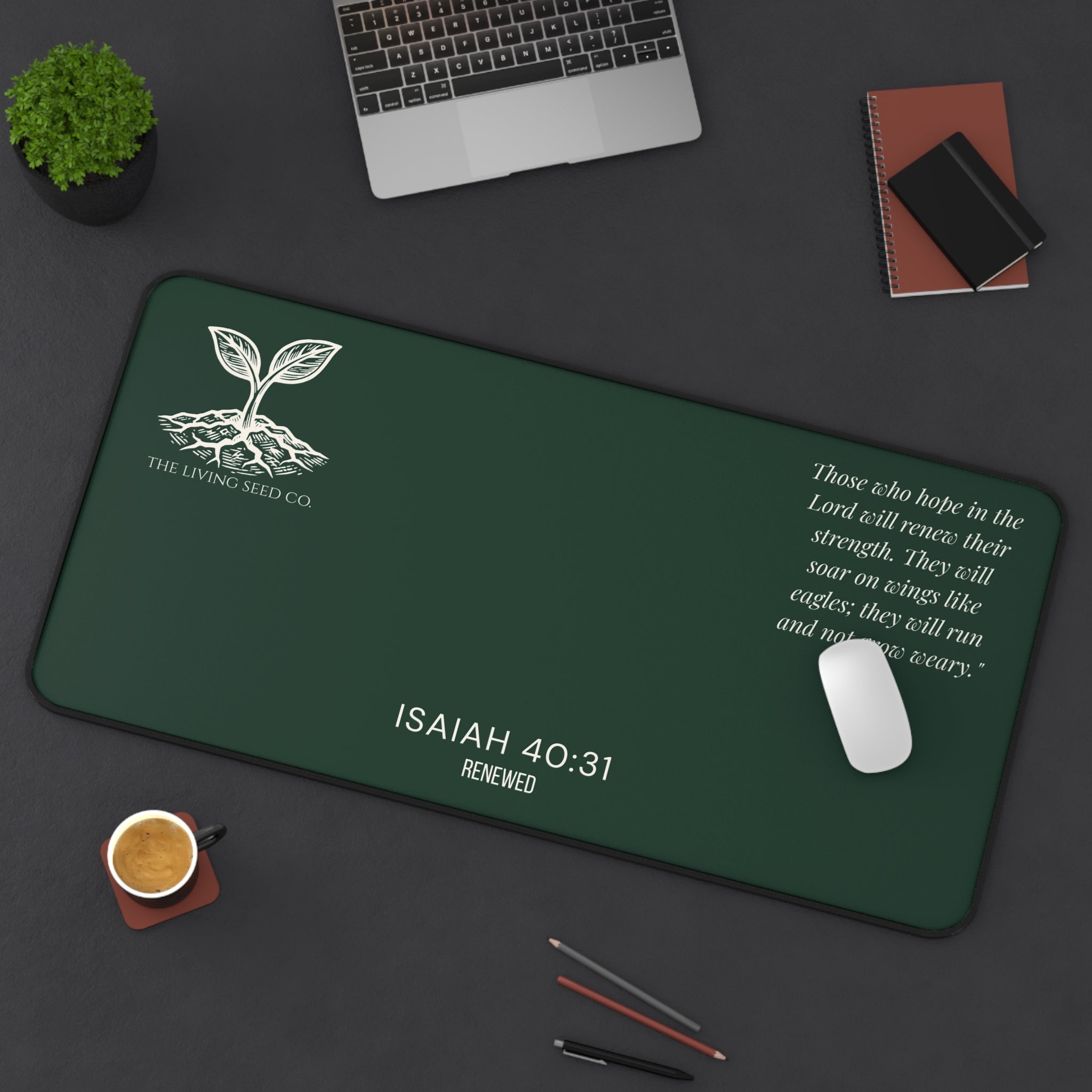 Scripture Series RENEWED Desk Mat in Forest Deep 16x32 — lifestyle shot — forest green desk mat on a dark workspace with laptop, coffee, and stationery featuring The Living Seed Co. seedling logo, Isaiah 40:31, RENEWED wordmark, and full Isaiah 40:31 verse in parchment ink.