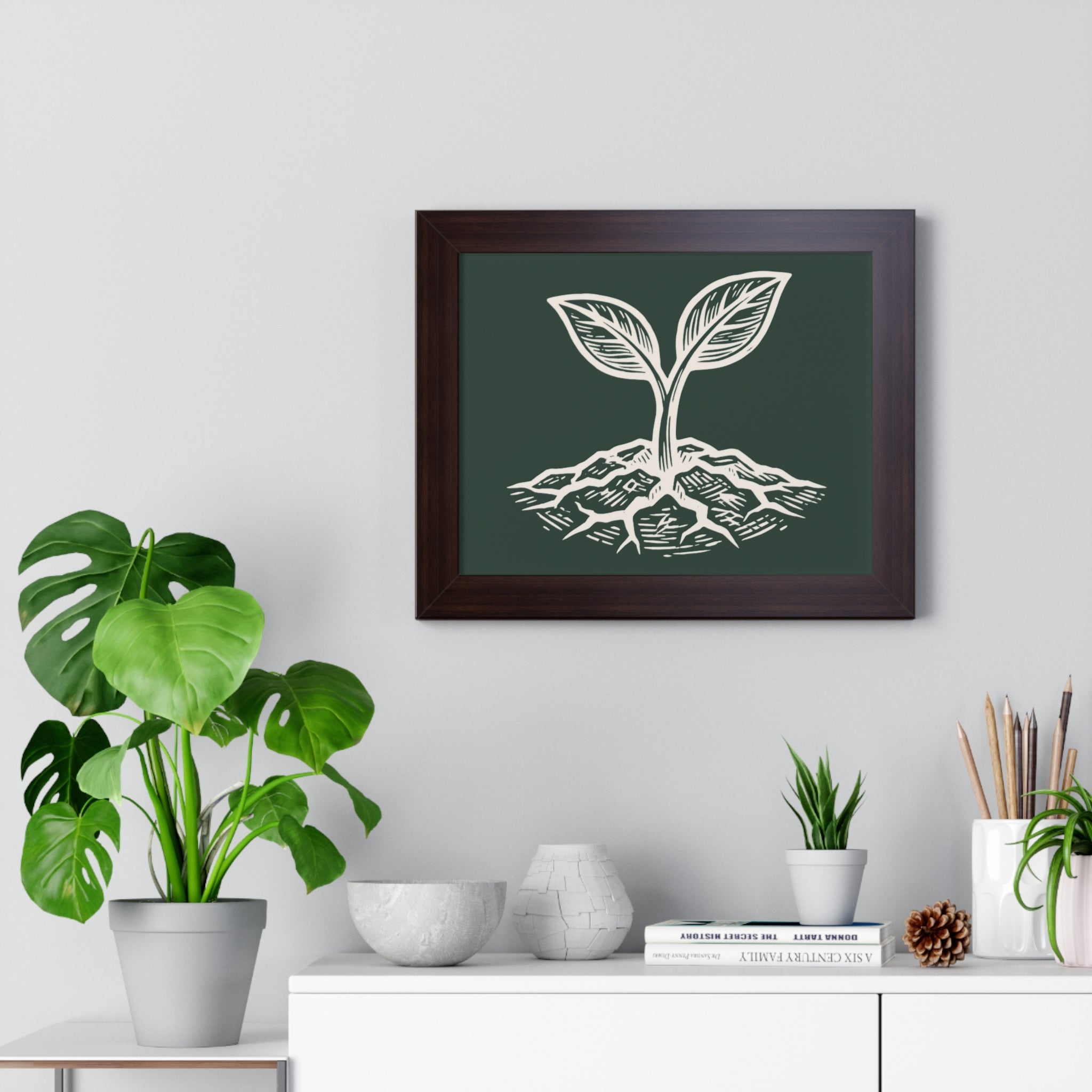 The Original Framed Poster in Walnut hanging on a gray wall above a shelf with houseplants, featuring an etching-style sprouting seed illustration on a Forest Deep background